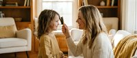 Young Caucasian mother and daughter sharing tender moment at home, woman gently touching child's face in cozy living room with warm natural lighting.