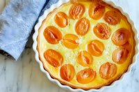 Fresh apricot custard tart with golden fruit arranged in ceramic baking dish on marble surface, homemade French dessert with bright orange halved apricots.