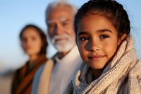 Heartwarming portrait of Hispanic multi-generational family outdoors at sunset, young girl in cozy knit scarf smiling at camera while standing with mother and grandfather.