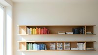 Modern wooden floating shelves with colorful books and magazines on white wall near window. Contemporary home library storage solution for living room.