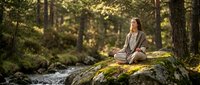 Young Caucasian woman practicing meditation in peaceful forest setting on moss-covered rock near flowing stream during golden hour sunlight.