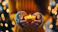 Female hands in cozy knitted sweater sleeves holding burning candle with golden bokeh lights background creating warm atmospheric winter mood.
