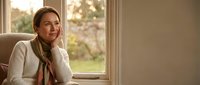 Middle-aged woman sitting by window in warm sunlight, hand on chin in thoughtful pose. Cozy home interior with natural lighting for lifestyle content.