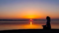 Woman meditating in lotus pose by calm lake at golden sunset with reflection on water creating peaceful zen atmosphere for wellness and mindfulness practice.
