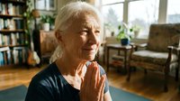 Senior Caucasian woman with white hair practicing yoga meditation at home, hands in prayer pose, peaceful expression in cozy living room.