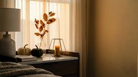 Cozy autumn bedroom interior with warm sunlight streaming through window, featuring decorative pumpkins and orange leaves on nightstand for seasonal home decor.