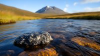 Tranquil mountain stream with dark rock emerging from flowing water, snow-capped peak in background, golden autumn grass on riverbank creates serene wilderness scene.