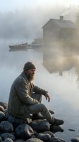 Bearded fisherman sitting on rocky shore at misty lake with wooden cabin and boat in background during golden hour sunrise.