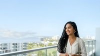 Young woman with long braided hair and South Asian ethnicity wearing a light beige blazer, standing on a high-rise balcony overlooking a cityscape. Professional business portrait.