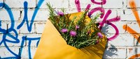 Yellow paper wrapped bouquet with wildflowers against colorful graffiti brick wall. Fresh floral arrangement for gifting, celebrations, urban lifestyle.