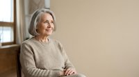 Senior woman with gray hair wearing beige sweater sitting indoors by window, looking away thoughtfully. Portrait for aging, lifestyle, wellness, retirement content.
