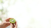Hand holding a sliced kiwi fruit with green flesh and seeds visible against a blurred natural background, suitable for food and nutrition content.
