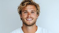 Young Caucasian man with wavy blonde hair and bright smile wearing white t-shirt against light background, looking directly at camera with confident expression.