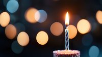 Single lit birthday candle on cupcake with sprinkles against dark background with soft glowing bokeh lights creating festive celebration atmosphere.