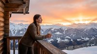 Woman in brown jacket holding warm beverage on snowy mountain cabin balcony at sunset, alpine landscape background, winter travel and relaxation concept.