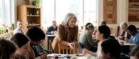 Mature Caucasian female teacher helping diverse students in bright classroom with wooden furniture and natural lighting for educational content.