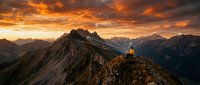 Hiker standing on mountain peak at dramatic golden sunset with alpine ridges and valleys below, suitable for travel and adventure marketing.