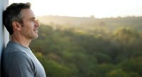 Middle-aged man with dark hair gazing at forested landscape from window, contemplative mood, nature view background.