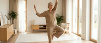 Senior man with gray hair, Caucasian ethnicity, performing yoga or stretching exercise in bright, minimalist living room with wooden furniture and plants for wellness and fitness content.