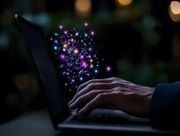 Magical sparkles and colorful light particles emerging from laptop keyboard while person types in dark environment with bokeh background effects.
