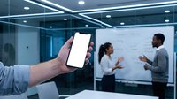 Hand holding blank white smartphone screen mockup in modern office with diverse business team presenting at whiteboard in background for app display.