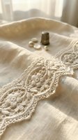 Cream linen fabric with ornate embroidered lace trim, metal thimble and scattered pearls on soft textile surface for sewing and crafts.