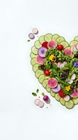 Heart-shaped salad arrangement with fresh cucumber slices, colorful radishes, cherry tomatoes, edible flowers and mixed greens on white background for healthy eating concepts.