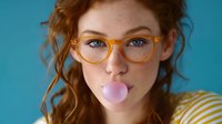 Young woman with ginger curly hair and yellow glasses blowing pink bubble gum against turquoise background, playful expression and casual style portrait.