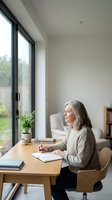 Mature woman with gray hair, Caucasian ethnicity, working at wooden desk near large black-framed windows, writing in notebook with pen, natural daylight streaming in, minimalist home office interior with potted plant, suitable for remote work, productivity, and lifestyle concepts.