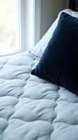 Cozy bedroom corner with navy blue pillow on light blue quilted bedding near bright window, creating serene morning atmosphere for relaxation and sleep design projects.