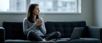 Young woman relaxing on couch with coffee and laptop in modern living room. Remote work, freelancing, home office lifestyle concept.