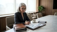 Senior businesswoman with gray hair and glasses reviewing financial documents at modern office desk with coffee cup and tablet.