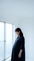 Pregnant Asian woman in dark blue dress standing by window in minimalist white room expecting baby maternal health wellness lifestyle.