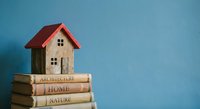 Miniature wooden house model with red roof sitting on stack of architecture and home design books against blue background for real estate concepts.