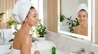 Young woman with towel on head applying skincare serum in modern bathroom mirror reflection for daily beauty routine and self-care wellness.