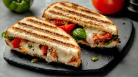 Grilled sandwich with melted mozzarella cheese, fresh tomatoes, and basil leaves on toasted bread, served on black stone board with vegetables in background.