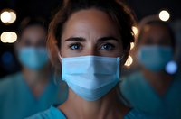 Female medical worker wearing protective face mask with determined look, colleagues in background. Healthcare frontline staff during pandemic crisis.