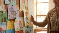Young woman admiring colorful children drawings pinned on a cork board in a bright room, showcasing creativity and learning.