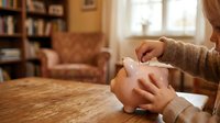 Caucasian child's hands placing coin into pink piggy bank on wooden table in cozy home library setting with bookshelf and armchair for savings and financial education concepts.