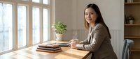 Young Asian businesswoman working at wooden desk in modern office with natural light and plants creating productive workspace atmosphere.