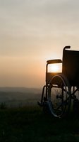 Wheelchair silhouetted against golden sunset sky on grassy hilltop overlooking misty valley landscape. Mobility aid symbolizing freedom, hope, and independence during golden hour.