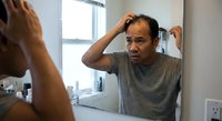 Middle-aged Asian man examining hair loss in bathroom mirror, touching scalp with concerned expression. Hair care and male pattern baldness concept.