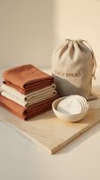 Folded linen kitchen towels in terracotta and beige tones with a drawstring storage bag and ceramic bowl on a wooden board.