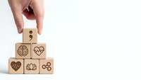 Hand placing wooden cube with semicolon symbol on pyramid of blocks featuring mental health icons including brain, heart, lotus flower on white background.