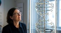 Middle-aged Caucasian female scientist in dark blazer examining large DNA double helix model in modern laboratory setting for genetic research and education.