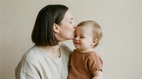 Young mother with dark hair tenderly kissing blonde toddler child on forehead in warm embrace against neutral beige background showing maternal love and bonding.