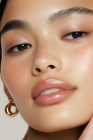 Close-up portrait of young Asian woman with natural freckles, dewy glowing skin, glossy coral lips, and gold hoop earring against neutral background.