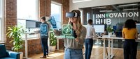 Young woman using VR headset in modern innovation hub office with diverse team collaborating on technology projects and digital interfaces.