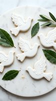White dove-shaped cookies with decorative icing arranged on marble surface with fresh green leaves for peaceful celebration or religious holiday baking.