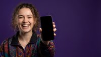 Happy young Caucasian woman with curly hair showing blank smartphone screen on purple background for mobile app presentation and digital marketing.